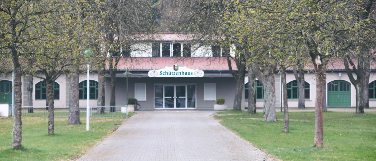 Breit angelegter, gepflasterter Weg führt zum historischen Schützenhaus, umgeben von laubfreien Bäumen.
