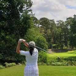 Golferin im weißen Outfit beim Abschlag auf einem von Wald umgebenen Golfplatz unter blauem Himmel.