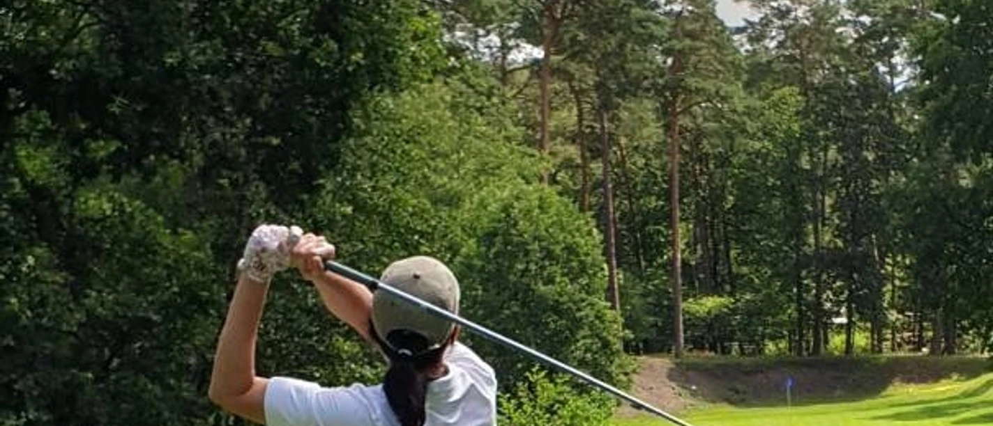 Golferin im weißen Outfit beim Abschlag auf einem von Wald umgebenen Golfplatz unter blauem Himmel.