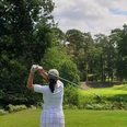 Golf.jpg Golferin im weißen Outfit beim Abschlag auf einem von Wald umgebenen Golfplatz unter blauem Himmel.