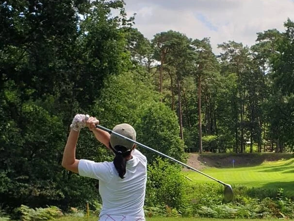 Golf.jpg Golferin im weißen Outfit beim Abschlag auf einem von Wald umgebenen Golfplatz unter blauem Himmel.