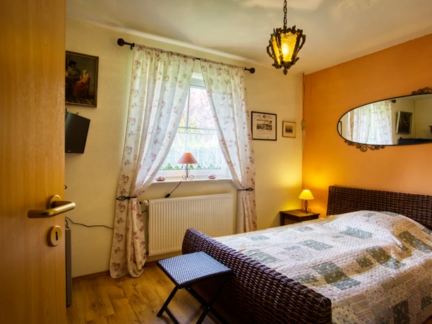 Schlafzimmer Doppelzimmer mit Rattanbett, Spiegel an orangefarbener Wand, warmer Beleuchtung und Fenster.
