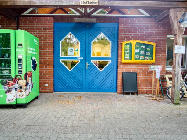 Hofladen Eingang eines Hofladens aus rotem Backstein mit blauer Holztür und grünem Regio-Automaten davor.