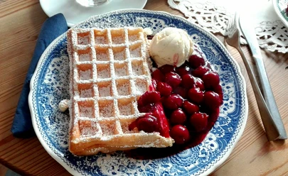 Waffel mit Puderzucker, Kirschen und Vanilleeis auf blauem Porzellanteller, auf Holzuntergrund.