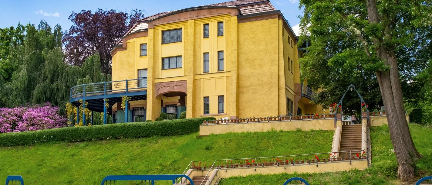KSC-Villa Esche-Foto-FrankKrueger-entsättigt-m.jpg Historische Villa mit gelber Fassade und Treppenzugang, umgeben von Garten und Bäumen.