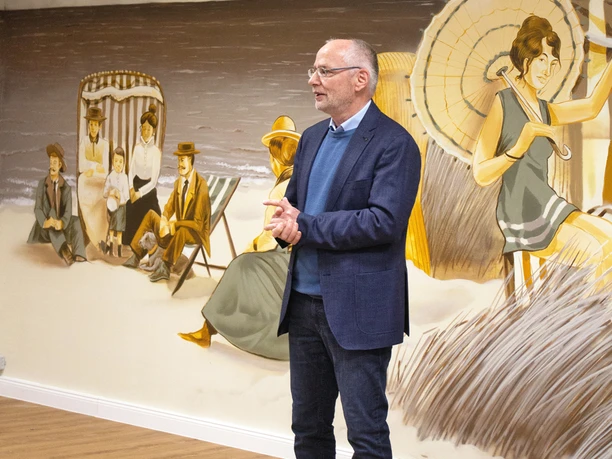 Strandkorbgeflüster mit Enno Ein Mann steht lächelnd und gestikulierend vor einer Wand mit einem nostalgischen Strandgemälde.