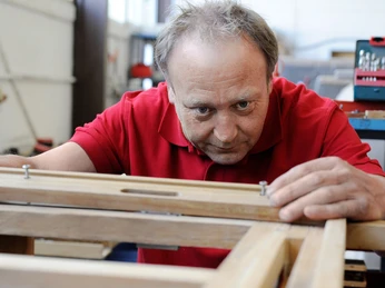 Holzarbeiten bei der Strandkorbherstellung Ein Angestellter im roten Polohemd arbeitet konzentriert an einem Holzrahmen in einer Werkstatt für Strandkörbe. Er justiert präzise die Befestigungen der Holzteile.
