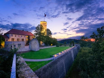 Bielefeld-Sparrenburg-blau-Wienke_Baustein und Teaser.jpg Burg mit rundem Turm und dahinter idyllischem Sonnenuntergang, umgeben von üppigem Grün und Mauer.