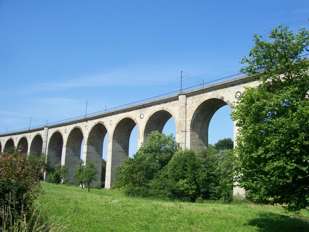 Steinviadukt mit Bögen und Gleisen, eingebettet in grüne Landschaft, blauer Himmel im Hintergrund.