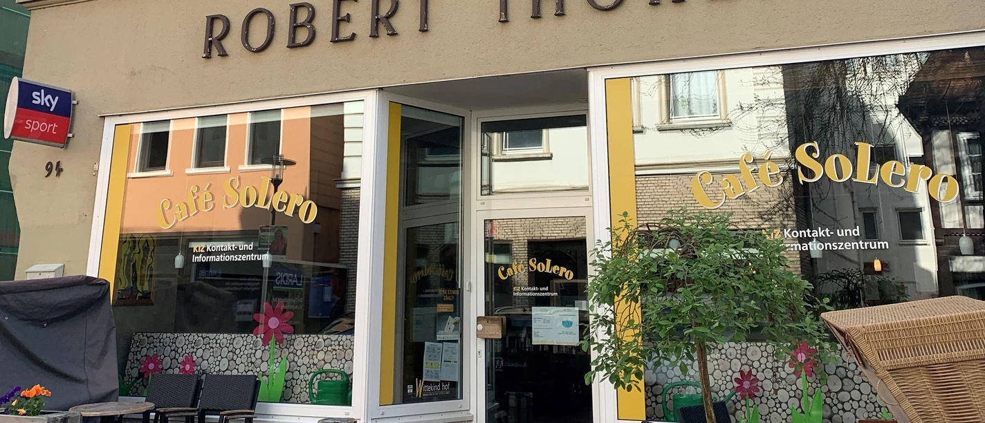 Eingang des Café Solero mit Sitzbereich im Freien. Gelbe Markisen und Blumen geben dem Café ein lebendiges Aussehen.