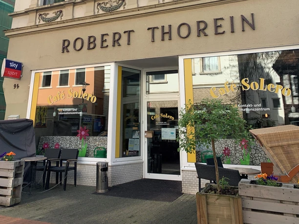 Außenansicht Cafe Solero Eingang des Café Solero mit Sitzbereich im Freien. Gelbe Markisen und Blumen geben dem Café ein lebendiges Aussehen.