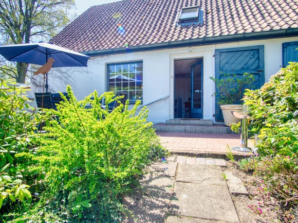 Weiße Fassade mit blauen Fensterläden und rotem Ziegeldach, umgeben von blühenden Sträuchern und Gartenpflanzen. Vor der Tür eine kleine Terrasse mit Gartenmöbeln und Sonnenschirm, die in einen begrünten Außenbereich übergeht.