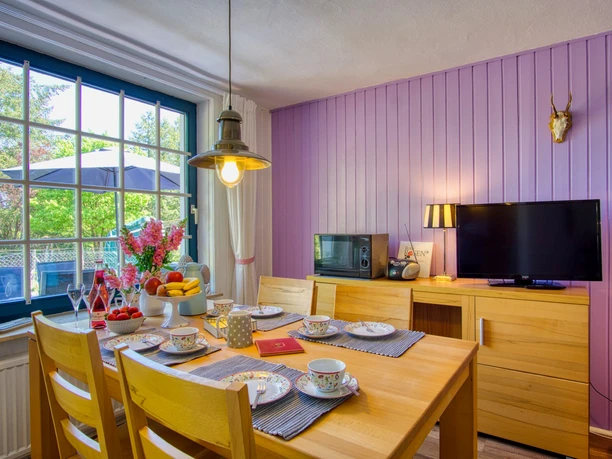 Heller Essbereich mit Holztisch und vier Stühlen, gedeckt mit buntem Geschirr und frischem Obst. Große Fenster mit Blick in den Garten, eine violett gestrichene Holzverkleidung an der Wand, Fernseher und Lampe schaffen eine freundliche Atmosphäre.
