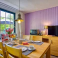 Willkommen! Heller Essbereich mit Holztisch und vier Stühlen, gedeckt mit buntem Geschirr und frischem Obst. Große Fenster mit Blick in den Garten, eine violett gestrichene Holzverkleidung an der Wand, Fernseher und Lampe schaffen eine freundliche Atmosphäre.