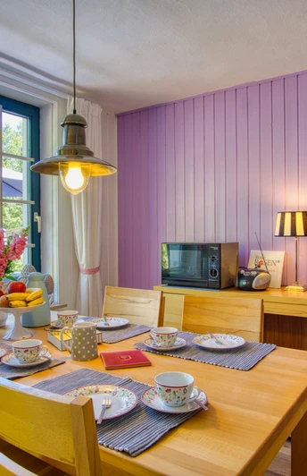 Willkommen! Heller Essbereich mit Holztisch und vier Stühlen, gedeckt mit buntem Geschirr und frischem Obst. Große Fenster mit Blick in den Garten, eine violett gestrichene Holzverkleidung an der Wand, Fernseher und Lampe schaffen eine freundliche Atmosphäre.