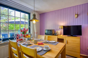 Willkommen! Heller Essbereich mit Holztisch und vier Stühlen, gedeckt mit buntem Geschirr und frischem Obst. Große Fenster mit Blick in den Garten, eine violett gestrichene Holzverkleidung an der Wand, Fernseher und Lampe schaffen eine freundliche Atmosphäre.