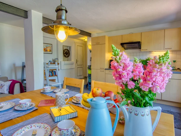 Helle Küche mit Holzfronten, integriertem Essbereich und liebevoll gedecktem Tisch mit floralem Geschirr, frischem Obst und pinken Blumen in einer Kanne. Im Hintergrund gemütlicher Wohnraum mit Sofa und Bücherecke.