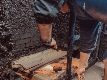 Ein Mann legt Holzscheite in einen Ofen, um Aale zu räuchern; Flammen leuchten im Hintergrund.