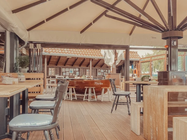 Schweer's Aalräucherei Schirmterrasse.jpg Eine mit Holzböden versehene Terrasse mit Stühlen und Tischgruppen unter einer großen Überdachung.