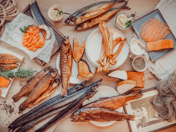 Schweer's Aalräucherei Räucherfisch.jpg Verschiedene Räucherfische wie Aal und Lachs liegen arrangiert auf einem Holztisch mit Dips und Kräutern.