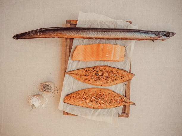 Schweer's Aalräucherei Räucherfischmix.jpg Verschiedene geräucherte Fischsorten, darunter Aal, Lachs und Makrele, dekorativ auf Holzbrett.