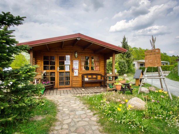 Rezeption Kleines Holzhaus mit rotem Dach und Blumenschmuck dient als Rezeption des Campingplatzes Lopautal. Ein gepflasterter Weg führt zum Eingang, umgeben von grüner Natur und bunten Blumen, die eine einladende Atmosphäre schaffen.