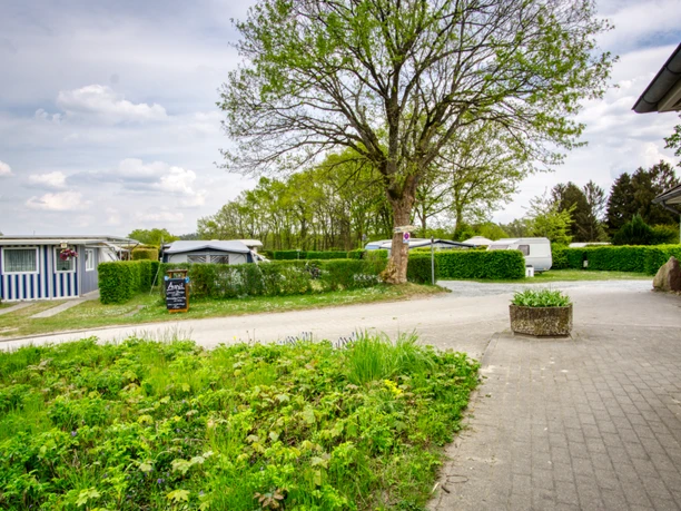 Campingplatz Lopautal Grüne Stellplätze mit Wohnwagen, gepflegten Wegen und hohen Bäumen auf einem weitläufigen Campinggelände. Im Vordergrund blühende Wiese und ein großes Schild vor einem kleinen Gebäude. Ruhige Umgebung mit Hecken und Natur im Hintergrund.
