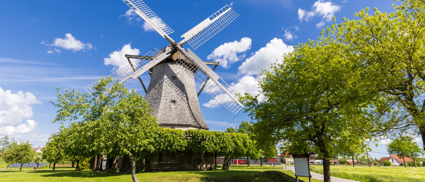Windmühle Destel Historische Windmühle mit rotierenden Flügeln unter blauem Himmel, umgeben von Bäumen und Wiesen.
