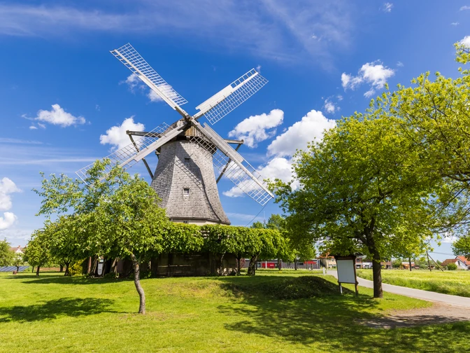 Windmühle Destel Historische Windmühle mit rotierenden Flügeln unter blauem Himmel, umgeben von Bäumen und Wiesen.