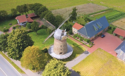 Historische Windmühle mit Steinstruktur und großen Flügeln, umgeben von grünen Feldern und Gebäuden.