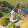 Historische Windmühle mit Steinstruktur und großen Flügeln, umgeben von grünen Feldern und Gebäuden.
