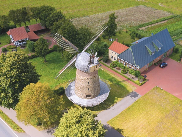 Historische Windmühle mit Steinstruktur und großen Flügeln, umgeben von grünen Feldern und Gebäuden.