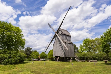Die historische Bockwindmühle Oppenwehe steht an grüner Wiese, umgeben von Bäumen und blauem Himmel.