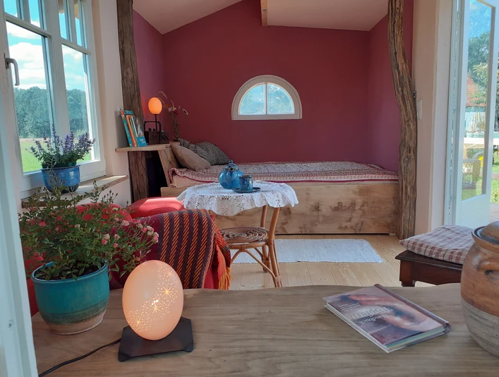 Bett im Tiny House Lille hus med træmøbler, vinduesudsigt til det grønne, træbord med lampe og magasin.