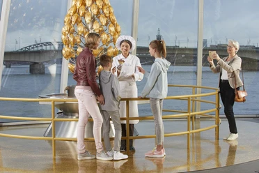 Schokoladenmuseum Im Schokoladenmuseum Köln führt eine Schokoladenexpertin in traditionellem Outfit eine Gruppe Kinder und Erwachsene durch die Ausstellung. Abgerundet wird die Szene durch eine moderne Kulisse mit Blick auf den Rhein.