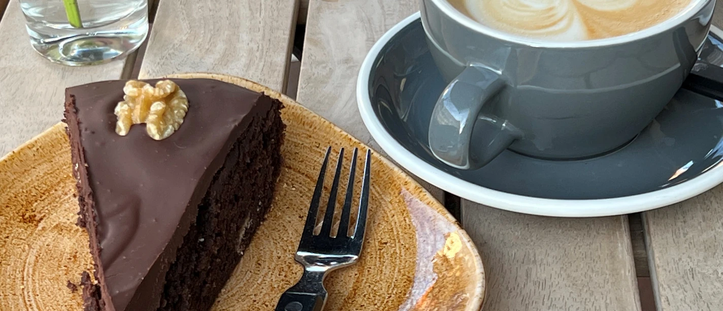 Een plak glazen chocoladetaart met walnoot op een bord naast een cappuccino met een hartpatroon.
