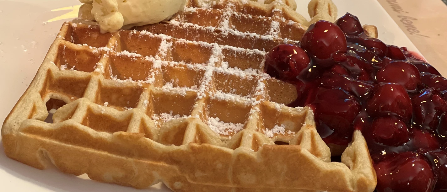 A freshly baked waffle with powdered sugar, cherries and a scoop of vanilla ice cream on a plate.