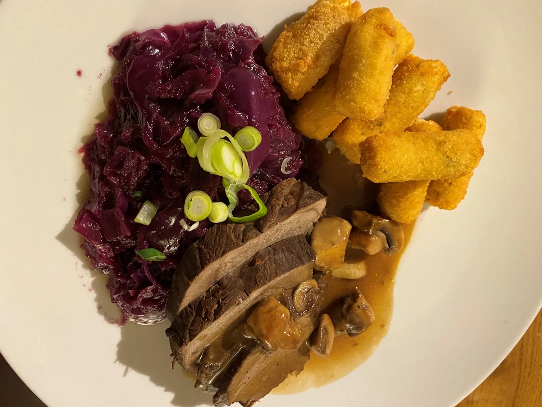 Hirschbraten mit glasierten Pilzen, serviert mit Rotkohl und knusprigen Kroketten auf weißem Teller.