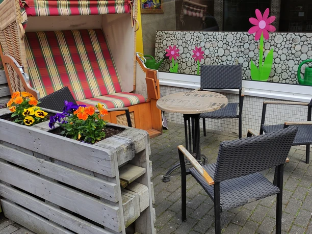 Cafe Solero Außenbereich Vier schwarze Rattan-Stühle um einen runden Holztisch vor einem floralen Schaufensterdekor.