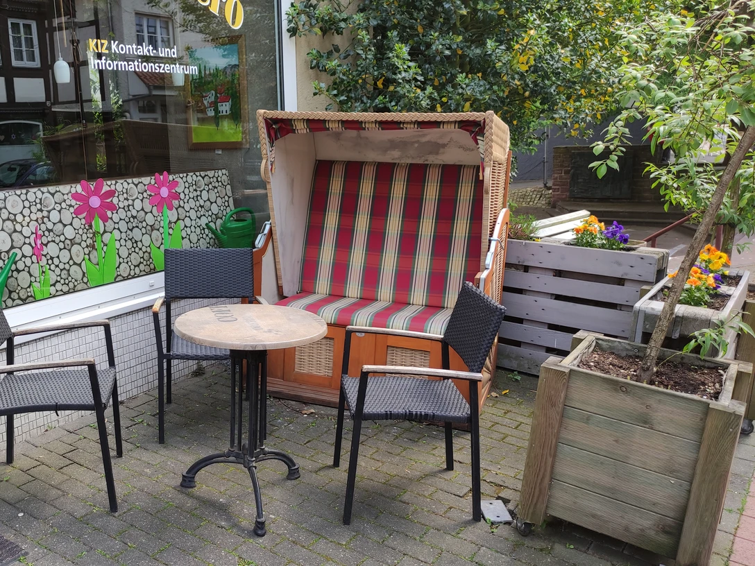 Drei Bistrostühle und ein runder Tisch stehen vor einem rot-weiß gestreiften Strandkorb auf gepflastertem Boden.