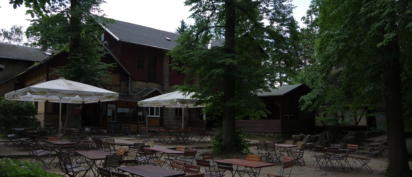 Berggaststätte Pfaffenstein Ein leerer Biergarten mit Holztischen und -stühlen unter großen Bäumen und Sonnenschirmen vor einem rustikalen Gebäude.
