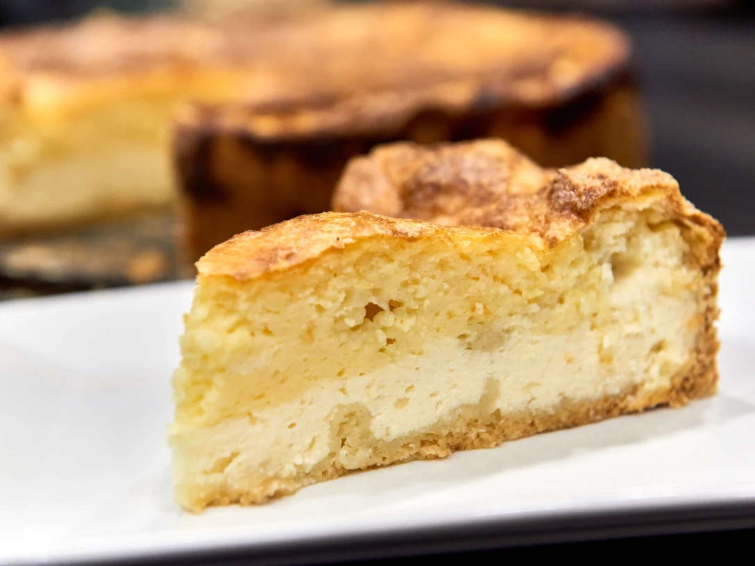 Ein Stück goldbrauner Käsekuchen auf einem weißen Teller, im Hintergrund der restliche Kuchen.A slice of golden brown cheesecake on a white plate, with the rest of the cake in the background.Kousek zlatohnědého tvarohového dortu na bílém talíři a zbytek dortu v pozadí.Kawałek złoto-brązowego sernika na białym talerzu, z resztą ciasta w tle.Een plak goudbruine cheesecake op een wit bord, met de rest van de taart op de achtergrond.Una fetta di cheesecake dorata su un piatto bianco, con il resto della torta sullo sfondo.