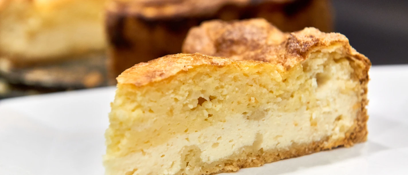 Een plak goudbruine cheesecake op een wit bord, met de rest van de taart op de achtergrond.