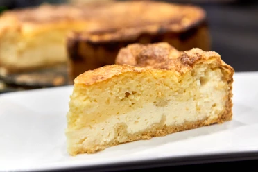 Wie macht man eine Eierschecke - Der typische Kuchen aus der Sächsischen Schweiz Ein Stück goldbrauner Käsekuchen auf einem weißen Teller, im Hintergrund der restliche Kuchen.