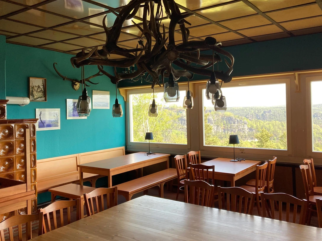Restaurace Felsgaststätte Rauenstein Gemütlicher Raum mit Holztischen und -stühlen, türkisfarbene Wände, Geweih-Lampen, große Fenster mit Blick auf bewaldete Landschaft.Cozy room with wooden tables and chairs, turquoise walls, antler lamps, large windows with a view of the wooded landscape.Útulná místnost s dřevěnými stoly a židlemi, tyrkysově zbarvené stěny, lampy z paroží, velká okna s výhledem na zalesněnou krajinu.Przytulny pokój z drewnianymi stołami i krzesłami, turkusowymi ścianami, lampami z poroża, dużymi oknami z widokiem na zalesiony krajobraz.Gezellige kamer met houten tafels en stoelen, turquoise gekleurde muren, lampen van gewei, grote ramen met uitzicht op het boslandschap.Sala accogliente con tavoli e sedie in legno, pareti color turchese, lampade in corno, ampie finestre con vista sul paesaggio boschivo.