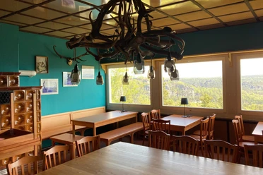 Restaurant Felsgaststätte Rauenstein Gezellige kamer met houten tafels en stoelen, turquoise gekleurde muren, lampen van gewei, grote ramen met uitzicht op het boslandschap.