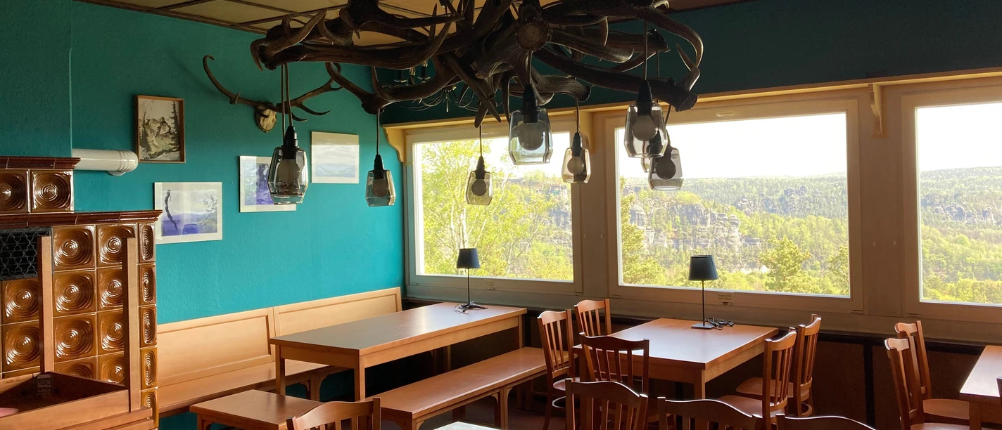 Restaurant Felsgaststätte Rauenstein Cozy room with wooden tables and chairs, turquoise walls, antler lamps, large windows with a view of the wooded landscape.