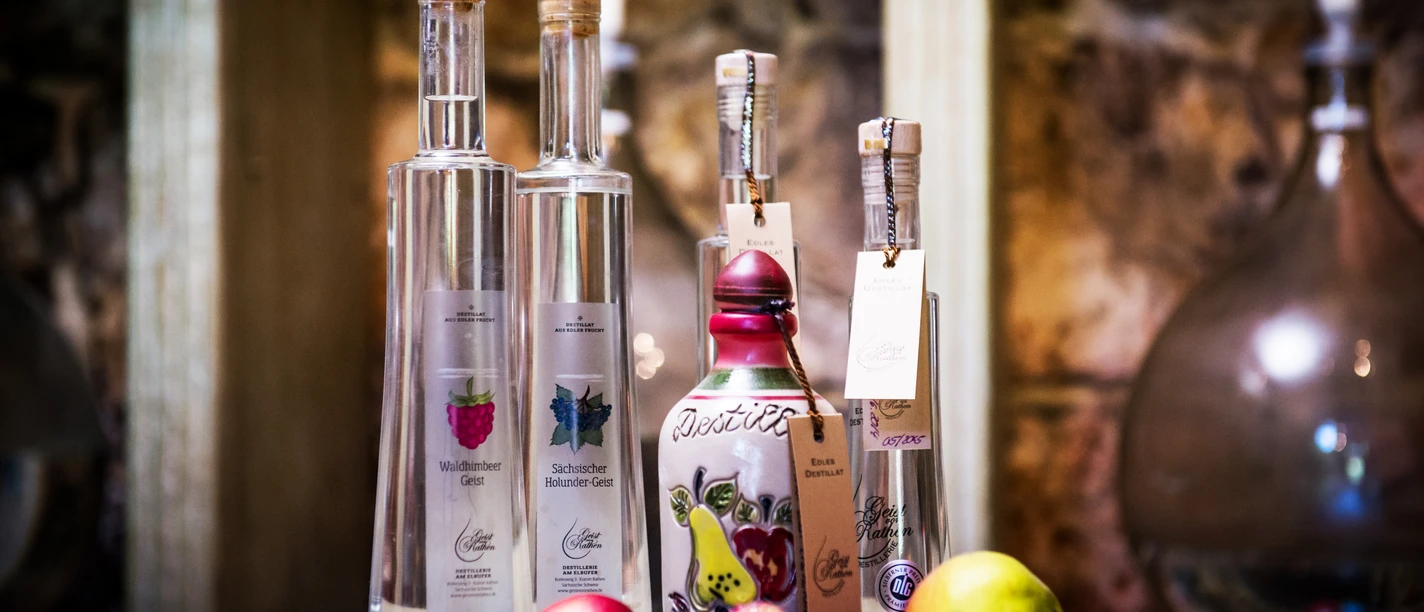 Pirna Distillery Various bottles of spirits on a table, surrounded by fresh fruit such as apples and pears, against a rustic backdrop.