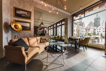 Restaurant Canaletto Gemütliches Café mit beiger Couch, schwarzem Kissen, rundem Glastisch und großen Fenstern mit Blick auf historische Gebäude.