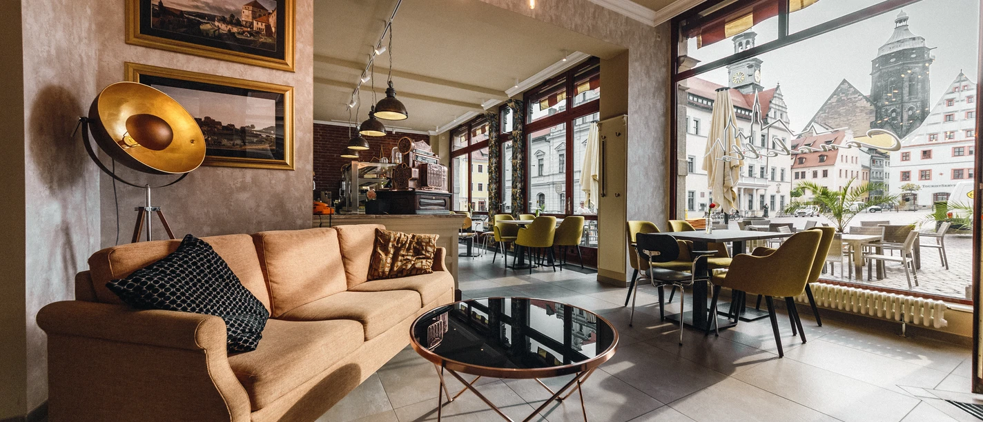 Restaurant Canaletto Gemütliches Café mit beiger Couch, schwarzem Kissen, rundem Glastisch und großen Fenstern mit Blick auf historische Gebäude.